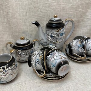 May include: A porcelain tea set featuring a teapot, a lidded sugar bowl, a creamer, and four teacups with saucers. Each piece has a black and white gradient finish with raised white dragon motifs, blue accents, and gold trim on the handles and rims.