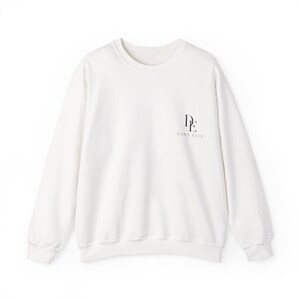Peut inclure: Un sweat-shirt à col rond et manches longues de couleur blanc cassé. L'avant présente un petit logo noir sur le côté supérieur droit composé des lettres DE empilées verticalement avec le texte DAWN ELVE imprimé en dessous.