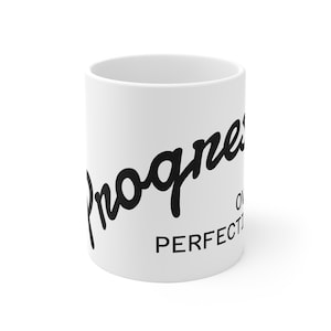 Peut inclure: Un mug en céramique blanche avec un texte noir indiquant Progress over perfection. Le mot Progress est écrit dans une grande police cursive grasse, tandis que les mots over perfection sont imprimés en dessous dans une police sans empattement.