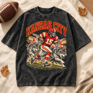 May include: A black acid-wash t-shirt featuring a graphic of a skeleton football player wearing a red and yellow jersey with the number 15, running alongside two wolves. The text KANSAS CITY is printed in large red and yellow arched letters.