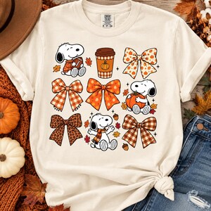 May include: A cream-colored short-sleeve t-shirt featuring a graphic print of Snoopy, various decorative bows in plaid and leopard patterns, a coffee cup with a pumpkin icon, and scattered autumn leaves and acorns in shades of orange and brown.