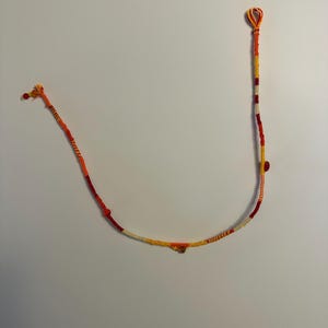 Puede incluir: Un collar de cordón largo y fino con una paleta de colores vibrantes de hilo rojo, naranja y amarillo envuelto alrededor de un núcleo central. El diseño incluye pequeñas cuentas dispersas en tonos cálidos y un lazo en el extremo.