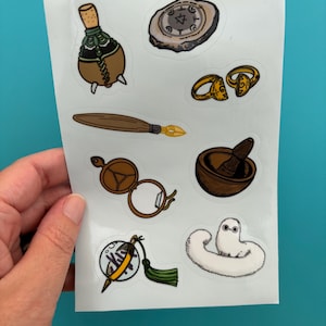 May include: A sticker sheet featuring various illustrated items including a brown potion bottle, a stone disc with symbols, a gold ring, a wooden quill, a brown locket, a mortar and pestle, a decorative charm with a green tassel, and a white fuzzy creature.
