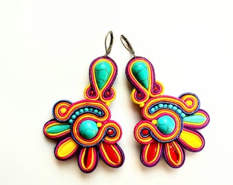 Puede incluir: Un par de pendientes coloridos con piedras turquesa y un diseño floral. Los pendientes están hechos de arcilla polimérica y tienen un esquema de colores amarillo brillante, rojo, rosa y morado.
