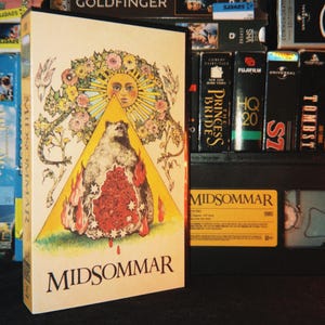 Peut inclure: Une cassette VHS du film Midsommar avec une couverture crème présentant un triangle jaune central contenant un ours entouré de fleurs rouges et de flammes, surmonté d'un soleil et d'une couronne florale, avec le titre Midsommar imprimé en bas.