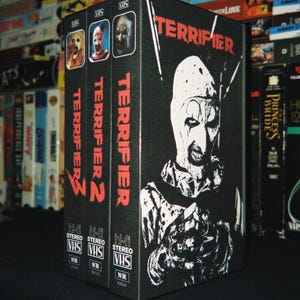 Può includere: Un cofanetto nero contenente tre videocassette VHS intitolate Terrifier, Terrifier 2 e Terrifier 3. La parte anteriore della scatola presenta una grafica bianca ad alto contrasto di un clown su sfondo nero con il testo rosso Terrifier.