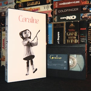 Könnte beinhalten: Eine weiße VHS-Hülle mit dem Titel Coraline in rosa Schreibschrift und einer schwarz-weißen Illustration einer Figur, die einen Stock hält, neben einer schwarzen VHS-Kassette mit einem Etikett, das den Titel Coraline in gelbem Text zeigt.