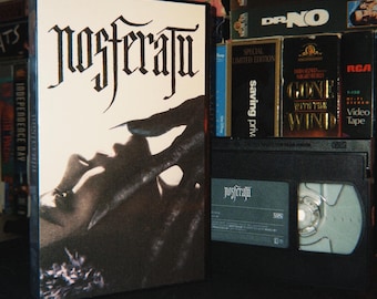 Könnte beinhalten: Eine Nosferatu VHS-Kassette in der originalen weißen Papphülle mit einer schwarz-weißen Grafik einer Hand, die nach einem Gesicht greift. Die schwarze Kunststoffkassette ist neben der Hülle zu sehen und zeigt den Titel Nosferatu in weißer Schrift.