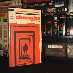 Könnte beinhalten: Eine VHS-Kassette mit dem Titel Obsession mit einem rot-schwarzen Cover, das die Silhouette einer Person in einer Reihe von ineinander verschachtelten Türen zeigt. Der Text lautet be careful who you wish for und obsession. Die Kassette steht zwischen anderen.