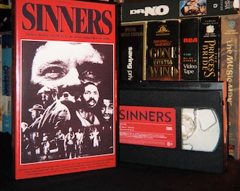 Könnte beinhalten: Eine Sinners VHS-Kassette mit rot-schwarzem Cover, das ein großes Gesicht und eine Gruppe von Menschen zeigt. Der Text lautet Sinners und You keep dancing with the devil, one day he's gonna follow you home. Die Kassette selbst ist schwarz.
