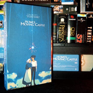 Könnte beinhalten: Eine blaue VHS-Kassette von Das wandelnde Schloss von Studio Ghibli mit einer Illustration von zwei Figuren, die unter einem hellen Stern in den Wolken stehen. Der Titel ist in weißer Schrift über den Figuren gedruckt, mit japanischem Text darüber.