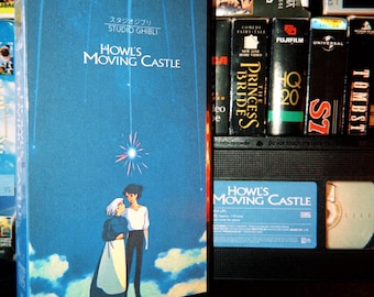 Könnte beinhalten: Eine blaue VHS-Kassette von Das wandelnde Schloss von Studio Ghibli mit einer Illustration von zwei Figuren, die unter einem hellen Stern in den Wolken stehen. Der Titel ist in weißer Schrift über den Figuren gedruckt, mit japanischem Text darüber.