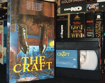 Könnte beinhalten: Eine VHS-Kassette des Films The Craft mit einem Cover, das den Text Light as a feather, stiff as a board und eine Grafik von zwei über einem Bett schwebenden Stiefeln zeigt. Die schwarze Kunststoffkassette steht neben der Hülle mit blauem Etikett.