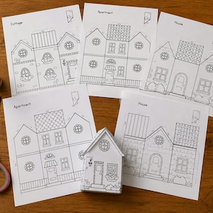 以下が含まれることがあります： Cottage、Apartment、Houseとラベル付けされたミニチュア構造物を組み立てるための黒い線画テンプレートが印刷された5枚の白い紙、組み立てられた小さな紙の家、スティックのり、ピンクの持ち手のハサミ。
