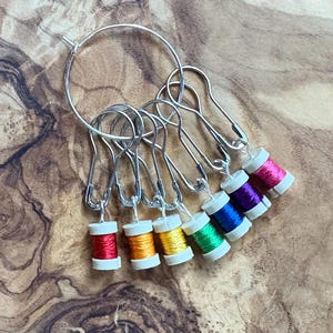 May include: A set of silver stitch markers with small spools of thread in various colors: red, orange, yellow, green, blue, purple, and pink. The stitch markers are attached to a silver hoop and are designed for knitting or crochet projects.