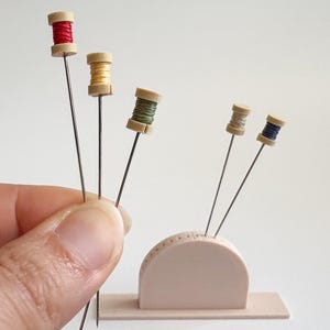 May include: A collection of sewing pins with miniature thread spools in red, yellow, and green. The pins are held by a hand and also displayed in a small, light pink pin holder. The spools have tan caps and are arranged in a visually appealing manner.