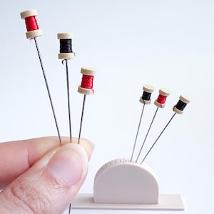 May include: Miniature sewing pins with red and black thread spools. The spools are mounted on silver pins and displayed in a small, light pink holder. The spools are made of beige and red or black thread.