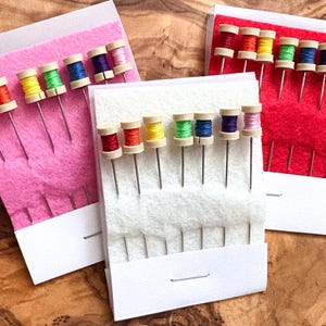 May include: Three sets of sewing needles with colorful thread spools. Each set has a different colored felt backing: pink, white, and red. The needles are arranged in a row, with the thread spools on top. The background is a wooden surface.