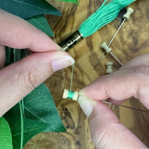 May include: Hands working with green embroidery floss, needles, and small wooden spools. The scene includes a skein of green thread and green leaves, suggesting a crafting or embroidery project. The wooden surface provides a natural backdrop.