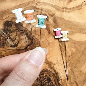May include: A collection of decorative sewing pins featuring miniature spools of thread in various colors: white, orange, teal, pink, and yellow. The spools are mounted on long, thin metal pins, ideal for crafting or adding a unique touch to sewing projects.