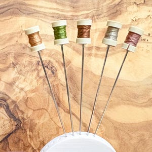 May include: Five colorful pin cushions with spools of thread in shades of brown, green, silver, and pink. Each spool is mounted on a long, thin metal pin. The pins are arranged in a fan shape against a wooden background.