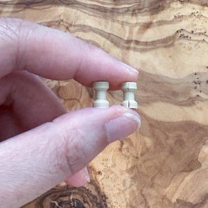 May include: Two small, light beige, cylindrical objects with a stepped design are held between fingers. The objects have a rounded top and bottom with a narrower middle section. The background is a wooden surface with a natural grain.