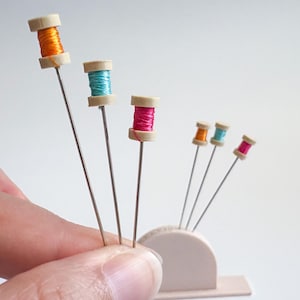 May include: A collection of colorful sewing pins with miniature spools of thread in orange, teal, and pink. The pins are held in a small, off-white holder, and some are held by a hand. The spools are attached to long, thin silver pins.