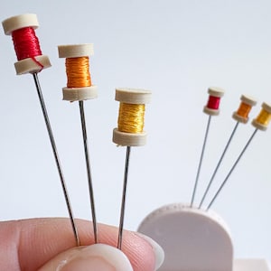 May include: A collection of miniature sewing pins with spools of thread in red, orange, and yellow. The spools are mounted on silver pins and are displayed against a white background. The pins are arranged in a decorative holder.