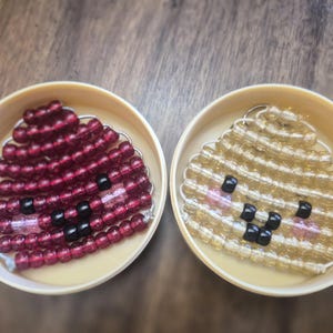 Peut inclure: Deux récipients circulaires contenant chacun un personnage en perles. Un personnage est fait de perles translucides rouge foncé avec des yeux et une bouche en perles noires. L'autre est fait de perles translucides or clair.