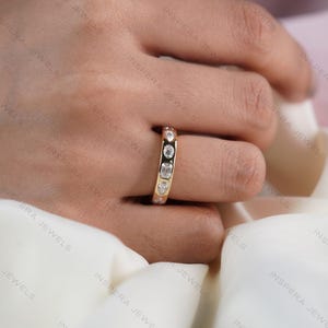 May include: A gold band ring featuring a series of bezel-set clear gemstones in various shapes, including round, emerald, and pear cuts, arranged along the center of the band. The ring has a polished finish and a smooth, rounded profile.