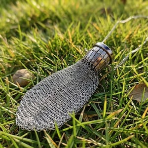 May include: A silver-toned chainmail pouch pendant featuring a metallic, cylindrical cap with vertical ridges and small rivets. The flexible, woven metal mesh body tapers to a rounded base, attached to a delicate silver-coloured metal chain.