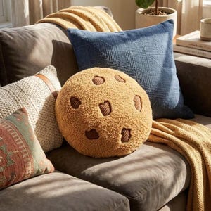 Peut inclure: Un coussin rond, beige et texturé en forme de biscuit aux pépites de chocolat, avec des accents en velours côtelé marron foncé. Le coussin est placé sur un canapé gris foncé à côté d'un coussin carré bleu texturé et d'un coussin en tricot crème.