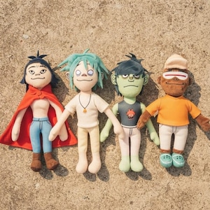 May include: Four plush dolls representing cartoon characters. The first wears a red cape, blue pants, and brown boots. The second has teal hair, a white shirt, and a necklace. The third is green-skinned with a dark tank top. The fourth wears an orange shirt, white pants, and a tan cap.