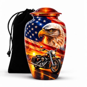 May include: A cylindrical metal cremation urn featuring a vibrant graphic of a bald eagle, the American flag, and a motorcycle on a road at sunset. The urn has a rounded lid and is accompanied by a black drawstring fabric storage bag.