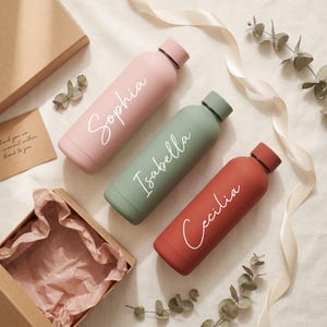 May include: Three cylindrical water bottles in matte pastel pink, sage green, and terracotta red. Each bottle features a white script name printed on the side: Sophia on the pink, Isabella on the green, and Cecilia on the red. Each has a matching cap.