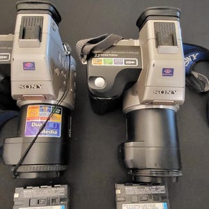 Puede incluir: Dos cámaras digitales Sony plateadas y negras con correas para el cuello y lentes negros. Cada cámara cuenta con una zapata para flash, una etiqueta de 2.3 Megapixel y una ranura Dual Media. Dos baterías negras rectangulares están debajo.