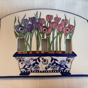 Könnte beinhalten: Eine dreifache Lichtschalterabdeckung aus Keramik mit weißem Hintergrund und blauem Rand. Das Design zeigt einen blau-weißen Pflanzkübel mit violetten und rosafarbenen Tulpen. Die Platte hat drei rechteckige Ausschnitte und sechs Schraublöcher.