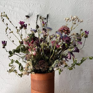 May include: A cylindrical terracotta vase with a ribbed texture containing a large arrangement of dried flowers in shades of purple, pink, and cream. The bouquet features various stems, delicate petals, and foliage extending outward from the top.