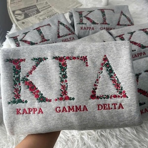 May include: A light gray sweatshirt featuring the Greek letters KΓΔ embroidered in a floral pattern with red flowers and green leaves. Below the letters, the words KAPPA GAMMA DELTA are embroidered in red thread. The fabric is a soft cotton blend.