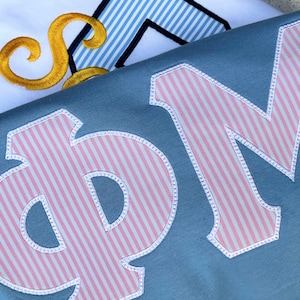 May include: A blue fabric garment featuring large Greek letters Phi and Mu appliqued in a pink and white vertical striped pattern with white stitched borders. In the background, a white fabric shows a gold embroidered script letter.