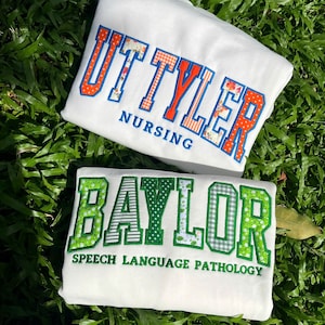 May include: Two white sweatshirts featuring embroidered collegiate-style lettering. The top sweatshirt displays 'UT TYLER' in orange patterned fabric with blue outlines and 'NURSING' in blue. The bottom sweatshirt shows 'BAYLOR' in green patterned fabric with green outlines and 'SPEECH LANGUAGE PATHOLOGY' in green.