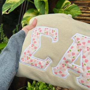 May include: A beige sweatshirt featuring large Greek letters Sigma and Alpha appliqued on the front. The letters are filled with a light pink fabric pattern of small cherries and strawberries, outlined with white embroidery stitching.