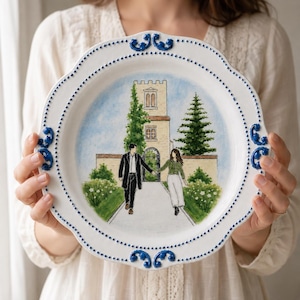 Puede incluir: Un plato de cerámica blanca con borde festoneado, detalles de cuentas azules y adornos en relieve de color azul. El centro muestra una escena pintada a mano de dos personas caminando de la mano por un sendero hacia un edificio de piedra con torre.
