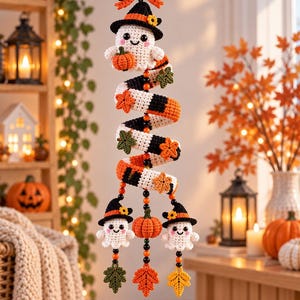 May include: A crochet hanging decoration featuring a spiral striped in orange, black, and white, adorned with small crocheted ghosts wearing black witch hats, orange pumpkins, and autumn leaves in green, orange, and yellow, finished with a bow.