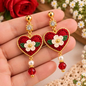 May include: A pair of gold-tone drop earrings featuring a red crocheted heart base with a central white flower and green leaves. Each earring is accented with a clear crystal stud, a white faux pearl, and a faceted red bead at the bottom.