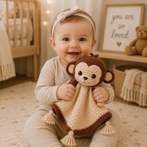 May include: A crochet monkey security blanket featuring a brown and beige colour scheme, a stuffed monkey head with embroidered facial features, pink cheeks, and four small tassels attached to the corners of the triangular crocheted body.