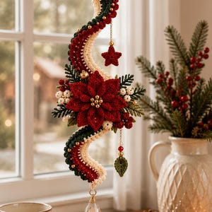 Op de afbeelding: Een hangend gehaakt ornament met een golvend, meerkleurig lint in crème, donkergroen en dieprood. Het is versierd met een centrale rode kerstster, kleine witte besjes, groene dennentakjes, een klein rood sterretje, een groen blaadje en een kristallen druppel.