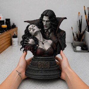 Op de afbeelding: Een gedetailleerd harsbeeld van een buste met Dracula met lang donker haar en een cape met hoge kraag die een vrouw met golvend haar in een donkerrood kanten lijfje omhelst. De voet heeft een gotisch ontwerp met het woord Dracula in reliëf.