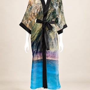 May include: A long-length robe featuring a photographic print of a rocky cliffside meeting blue water. The garment has black trim along the collar, cuffs, and front opening, and includes a matching black fabric tie belt at the waist.