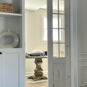 May include: A white door with a glass pane and a white trim. The door is open and you can see a wooden table with a white bowl of flowers on it.